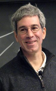A grey haired man is smiling shyly looking over your left shoulder